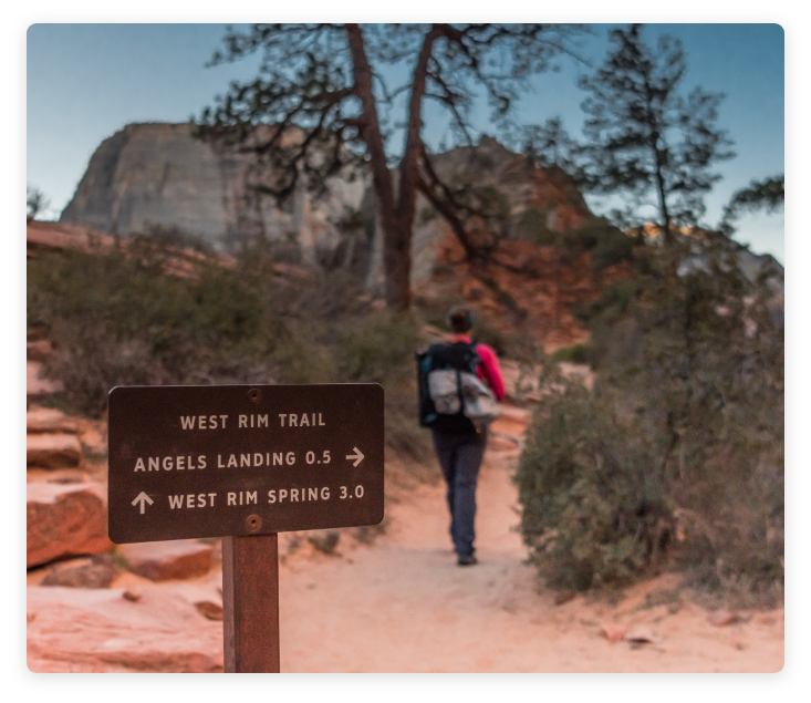 Individual with hiking gear hiking on trailhead in background with sign explaining trail information for "West Rim Train" in foreground.