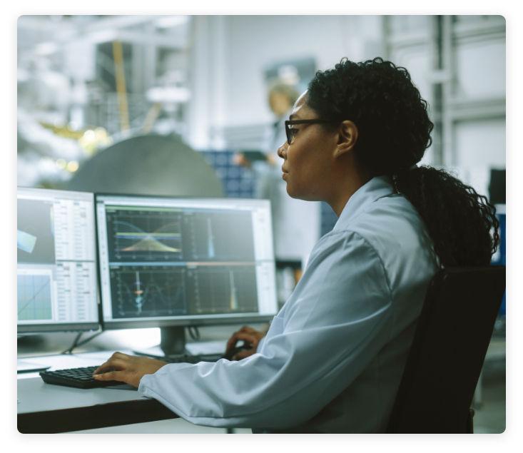 Female scientist in lab coat sitting computer with dual monitors, analyzing data on screens in laboratory.