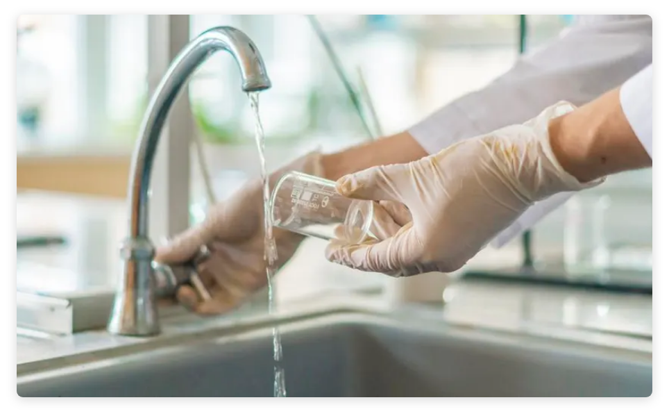 Detail of scientist wearing lab coat and rubber gloves while filling small glass beaker from running water faucet.