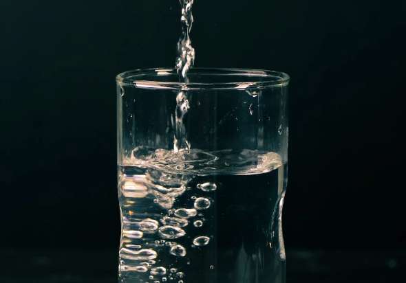 Water pouring into half full clear drinking glass over black background.
