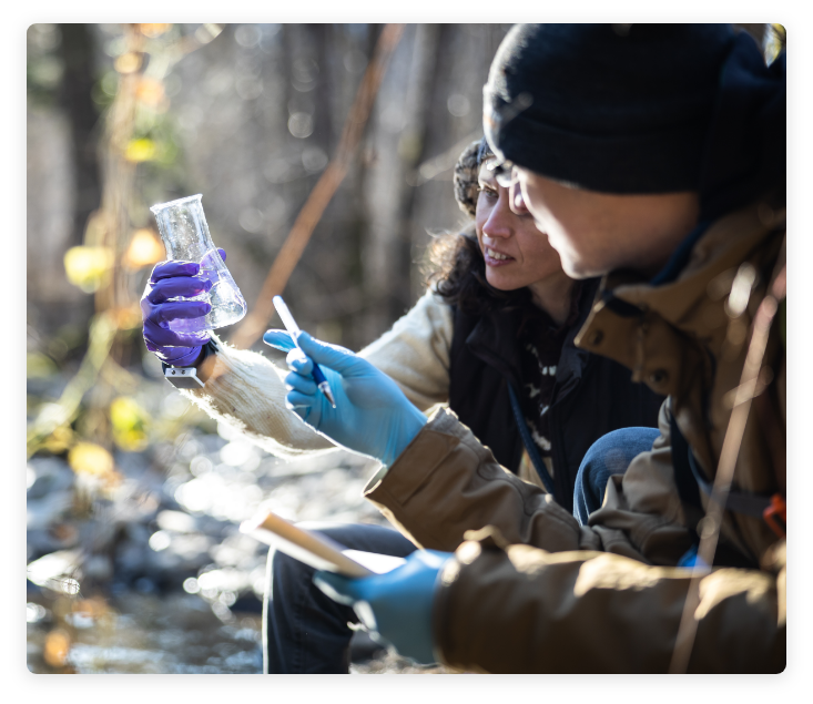 Detail of male and female in nature near water source, holding an Erlenmeyer flask and discussing its contents.