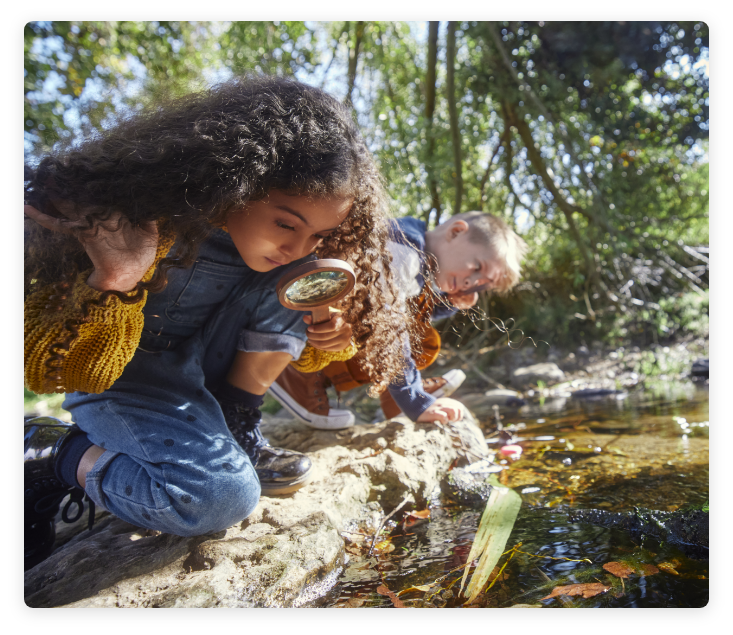 Children in nature near a water stream, holding magnifying glasses and analyzing contents of water.